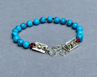 Op de afbeelding: Een armband met ronde turkoois kralen met donkere aders geregen aan een rood koord. Het bevat twee rechthoekige zilverkleurige metalen plaatjes, elk bezet met een kleine groene steen, een zilverkleurige karabijnsluiting en een ronde hanger met NB.