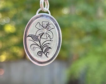 Op de afbeelding: Een ovale zilveren hanger met een gedetailleerd zwart gegraveerd bloemmotief en een kleine steen in het midden. De hanger hangt aan een zilveren ringetje en een metalen haak, met een gepolijste metalen afwerking.