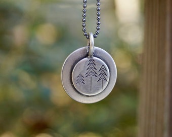 Op de afbeelding: Een zilveren ketting met een ronde hanger met een ontwerp van drie gestileerde bomen. De hanger is aan een zilveren ketting.
