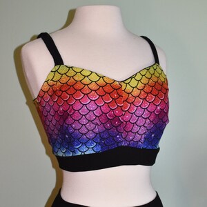 May include: A two-piece outfit with a rainbow mermaid scale print. The top is a cropped tank top with black straps and a black band at the bottom. The bottom is a pair of high-waisted shorts with a black waistband.
