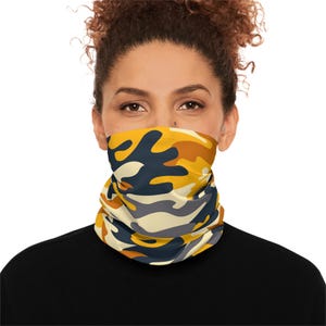 Könnte beinhalten: Ein gelber, blauer und grauer Camouflage-Halswärmer. Der Halswärmer wird von einer Person mit lockigem braunen Haar getragen.