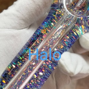 May include: A clear glass pipe with iridescent glitter embedded in the glass. The pipe has a rounded bowl and a long, straight stem. The glitter is a mix of blue, green, purple, and pink.