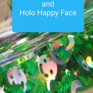 May include: Close-up of green and iridescent holographic glitter, featuring hemp leaf and happy face shapes. The glitter is contained in a clear container with the text "Holo Hemp Leaf and Holo Happy Face" on a blue background.