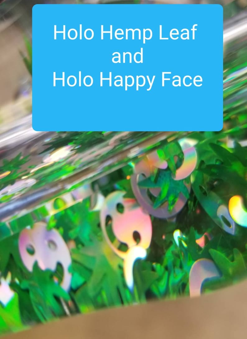 May include: Close-up of green and iridescent holographic glitter, featuring hemp leaf and happy face shapes. The glitter is contained in a clear container with the text "Holo Hemp Leaf and Holo Happy Face" on a blue background.