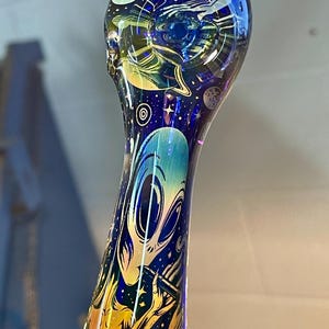 May include: A glass pipe with a blue, green, and purple iridescent finish. The pipe is decorated with an alien playing a flute in front of a starry night sky.