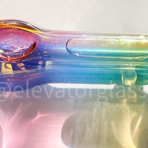 May include: A colorful glass pipe with a rainbow iridescent finish. The pipe features a bowl, a carb, and a mouthpiece. The colors transition from orange to blue and green. The text "@elevatorglass" is visible.