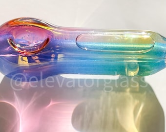 May include: A colorful glass pipe with a rainbow iridescent finish. The pipe features a bowl, a carb, and a mouthpiece. The colors transition from orange to blue and green. The text "@elevatorglass" is visible.