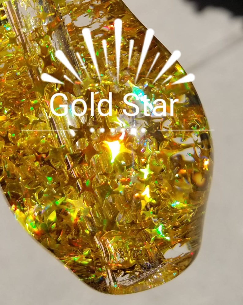 May include: A clear glass pipe with a gold glitter design. The pipe has a round shape and a small opening at the top. The glitter is iridescent and creates a shimmering effect. The text "Gold Star" is visible on the pipe.