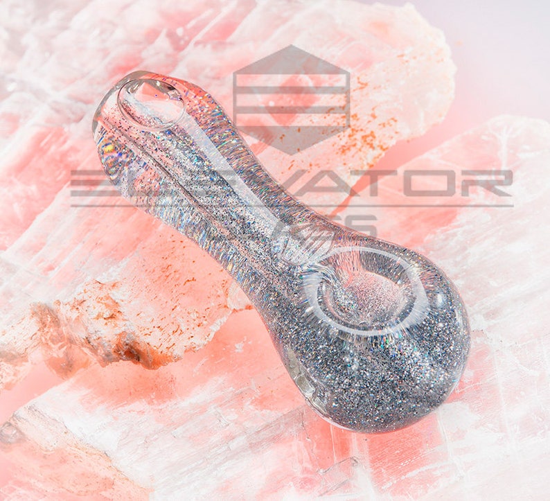 May include: A clear glass pipe with silver glitter embedded in the glass. The pipe is resting on a pink surface.