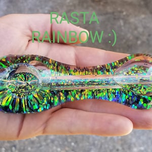 May include: A green and gold glass pipe with glitter embedded in the glass. The pipe is shaped like a spoon with a round bowl at one end and a long, thin stem at the other. The text "RASTA RAINBOW :)" is written in green letters on the image.