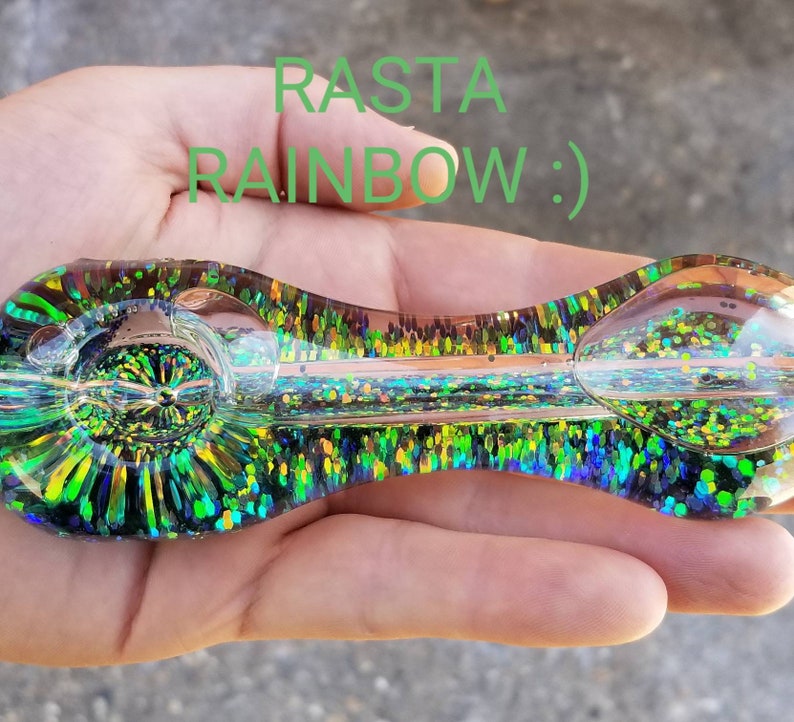 May include: A green and gold glass pipe with glitter embedded in the glass. The pipe is shaped like a spoon with a round bowl at one end and a long, thin stem at the other. The text "RASTA RAINBOW :)" is written in green letters on the image.