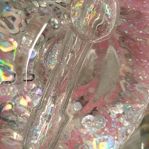 May include: Close-up of a clear glass object filled with pink glitter and iridescent hearts. The object has a clear glass rod running through it, and a large clear bubble at the top.