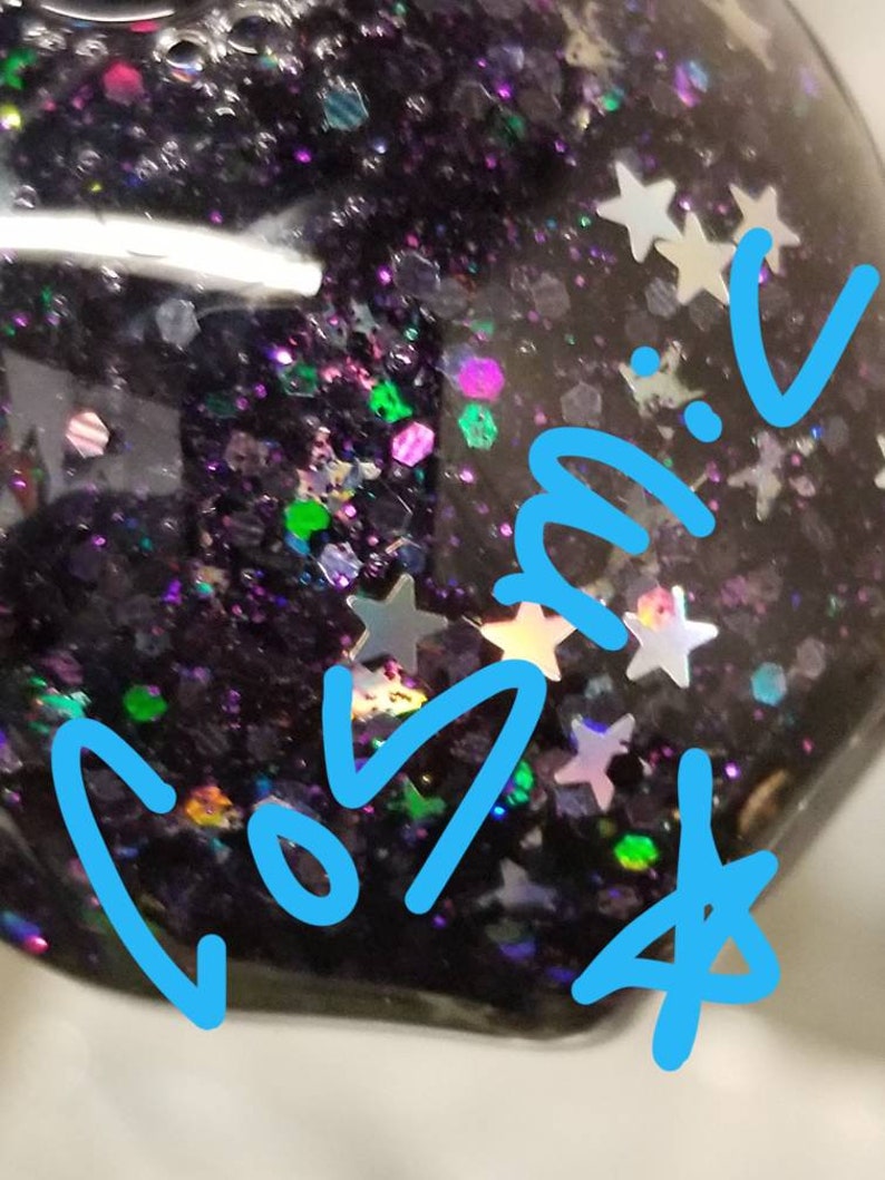 May include: A close-up of a black glitter nail polish with iridescent glitter and the word "COSMIC" written in blue on the bottle.