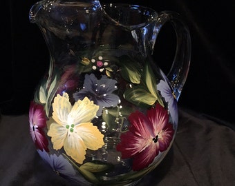 May include: A clear glass pitcher with a hand-painted design of purple, yellow, and red flowers. The flowers are painted in a realistic style and have green leaves.