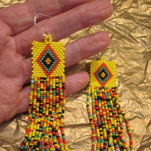 May include: A pair of long beaded earrings with a geometric pattern in yellow, red, green, and black. The earrings are made of small seed beads and have a fringe-like design.