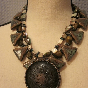 May include: A multi-strand necklace with brown, green, and white ceramic and gemstone beads. The necklace features a large, round, silver-toned pendant with an engraved design.