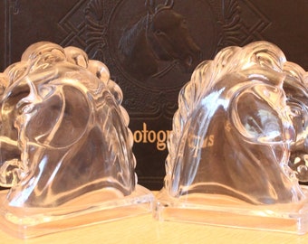 May include: A pair of clear glass horse head bookends. The bookends are sitting on a brown surface.