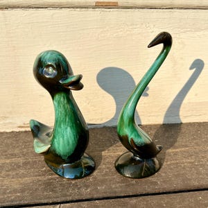 May include: Two ceramic duck figurines. One duck is upright with a long neck and the other is a duck with a shorter neck. Both are glazed in shades of green and brown, with a glossy finish. The figurines are placed on a wooden surface.
