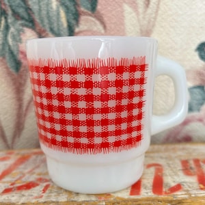 May include: A white milk glass mug featuring a prominent red and white checkered pattern that wraps around the center of the cylindrical body. The mug has a smooth, glossy finish, a sturdy side handle, and a slightly tapered base design.