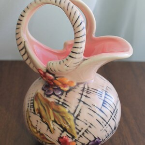 May include: A pink ceramic pitcher with a woven design and floral accents. The pitcher has a handle shaped like a basket.