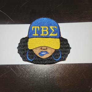 May include: Embroidered patch featuring a stylized Black woman wearing a blue and yellow baseball cap with the letters TBS in gold. The woman has blue hoop earrings, blue lipstick, and black curly hair. The patch is on a black and white striped background.
