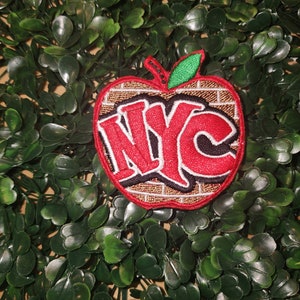 Può includere: Una mela rossa con un motivo a mattoni e la scritta "NYC" in lettere bianche e nere. La mela ha una foglia verde in cima.