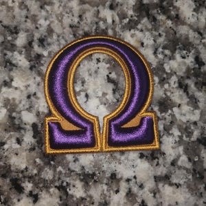 May include: Purple and gold embroidered iron-on patch with the Greek letter Omega.