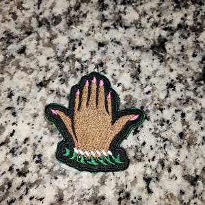 May include: A brown hand with pink fingernails is shown with a green leafy design around the wrist. The hand is raised with fingers extended.