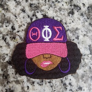 May include: Embroidered patch featuring a black woman with purple and pink hat, the letters "ΦΟΣ" on the hat, and large purple hoop earrings.