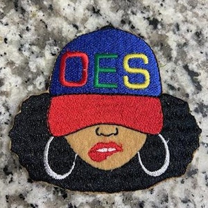 May include: Embroidered patch featuring a cartoon woman with an afro hairstyle wearing a blue, red, and yellow baseball cap with the letters "OES" on the front.