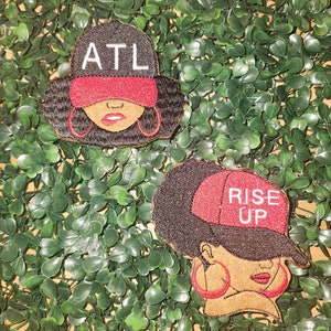 May include: Embroidered patches featuring stylized faces wearing baseball caps. One patch reads "ATL" in white letters on a black cap with a red visor. The other patch says "RISE UP" on a red cap.