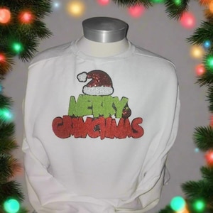 Puede incluir: Sudadera blanca de manga larga con el texto "MERRY GRINCHMAS" en purpurina verde y roja. El diseño incluye un gorro de Papá Noel. La sudadera se muestra en un maniquí con un árbol de Navidad y luces en el fondo.