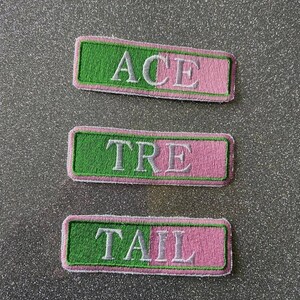 May include: Three embroidered patches with white lettering on a green and pink background. The patches read "ACE", "TRE", and "TAIL".