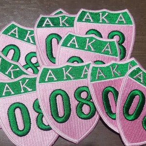 May include: A close-up of several pink and green embroidered patches with the letters "AKA" and the number "08" on them.