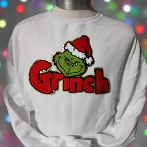 May include: White long-sleeved sweatshirt with a red and gold embroidered Grinch design. The Grinch is wearing a Santa hat and the word "Grinch" is written in gold letters.