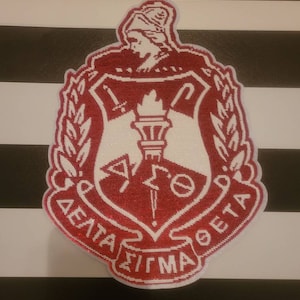 May include: A red and white embroidered patch with a crest featuring a woman's head, a torch, and a sword. The text "DELTA SIGMA THETA" is written around the crest.