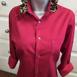 May include: A long-sleeved button-down shirt in a solid deep red color. The shirt features a distinctive ruffled collar with a brown and black leopard print pattern. It includes a single chest pocket on the left side and matching red buttons.