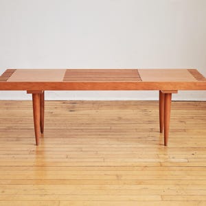 May include: A mid-century modern wooden bench with a natural finish. The bench features a slatted top and tapered legs. The wood has a warm, reddish-brown hue. The bench is approximately 60 inches long.
