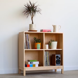 May include: A light wood bookshelf with four shelves, each with a different item. The top shelf has a white pitcher and two books. The second shelf has a potted plant and a stack of vinyl records. The third shelf has a potted plant and a white ceramic vase. The bottom shelf has a stack of books and a small speaker.
