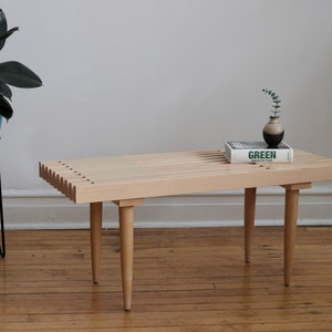 May include: A light wood bench with four legs and a slatted top. A book titled "GREEN ARCHITECTURE" with a small vase of greenery sits on top of the bench.