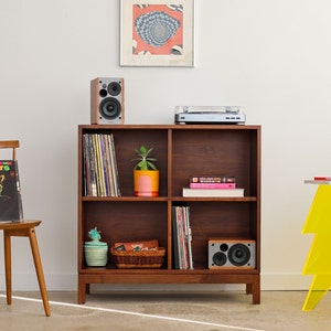 May include: A wooden bookshelf with four shelves, filled with vinyl records, books, and a potted plant. A record player sits on top of the bookshelf, and two speakers are on either side. A yellow lightning bolt shaped side table is on the right side of the image.