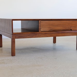 May include: A rectangular wooden coffee table with a warm brown finish. The table features a drawer on the right side and an open shelf on the left. It stands on four sturdy legs, showcasing a mid-century modern design.