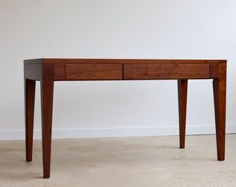 May include: A dark brown wooden desk with two drawers and tapered legs. The desk is sitting on a light brown carpet.