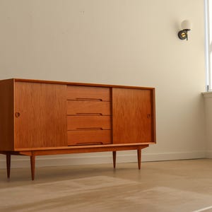 May include: A mid-century modern wooden sideboard with a warm, reddish-brown finish. The cabinet features sliding doors on either side and a set of three drawers in the center. It stands on tapered legs, offering a blend of storage and style.