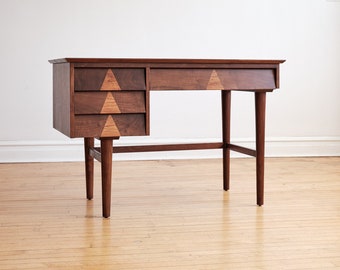 May include: A mid-century modern walnut desk with three drawers and a geometric inlay design. The desk has a simple, elegant design with tapered legs.