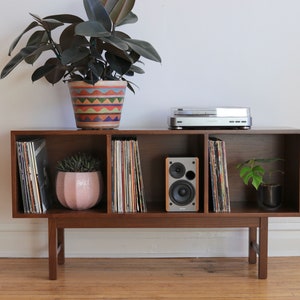 May include: A wooden record player stand with four shelves, two speakers, and a turntable. The stand has a light wood finish and is decorated with plants in colorful pots.