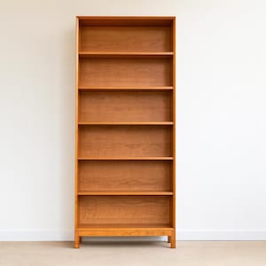 May include: A tall, wooden bookshelf with six shelves. The bookshelf is made of light brown wood and stands against a white wall. The shelves are evenly spaced, providing ample storage space for books and decorative items.