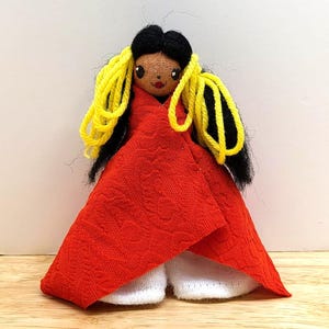 May include: A handmade doll with black hair and yellow yarn accents. The doll is wearing a red textured fabric dress and white shoes. The doll has a painted face with brown eyes and a red mouth.