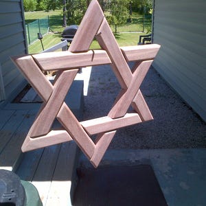 May include: A six-pointed Star of David constructed from smooth, light brown natural wood. The geometric design features interlocking straight wooden beams with visible grain patterns, creating a three-dimensional effect in this star-shaped object.