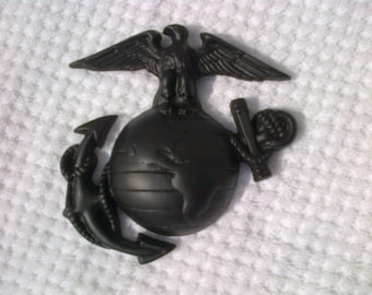 May include: A black metal eagle, globe, and anchor emblem. The eagle is perched on top of the globe, with its wings spread out. The anchor is below the globe, with a rope wrapped around it.